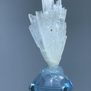 May include: A translucent, white, fan-shaped mineral cluster with vertical striations and a small brown inclusion, mounted atop a faceted, light blue, diamond-shaped glass base held by a silver-toned metal prong setting.