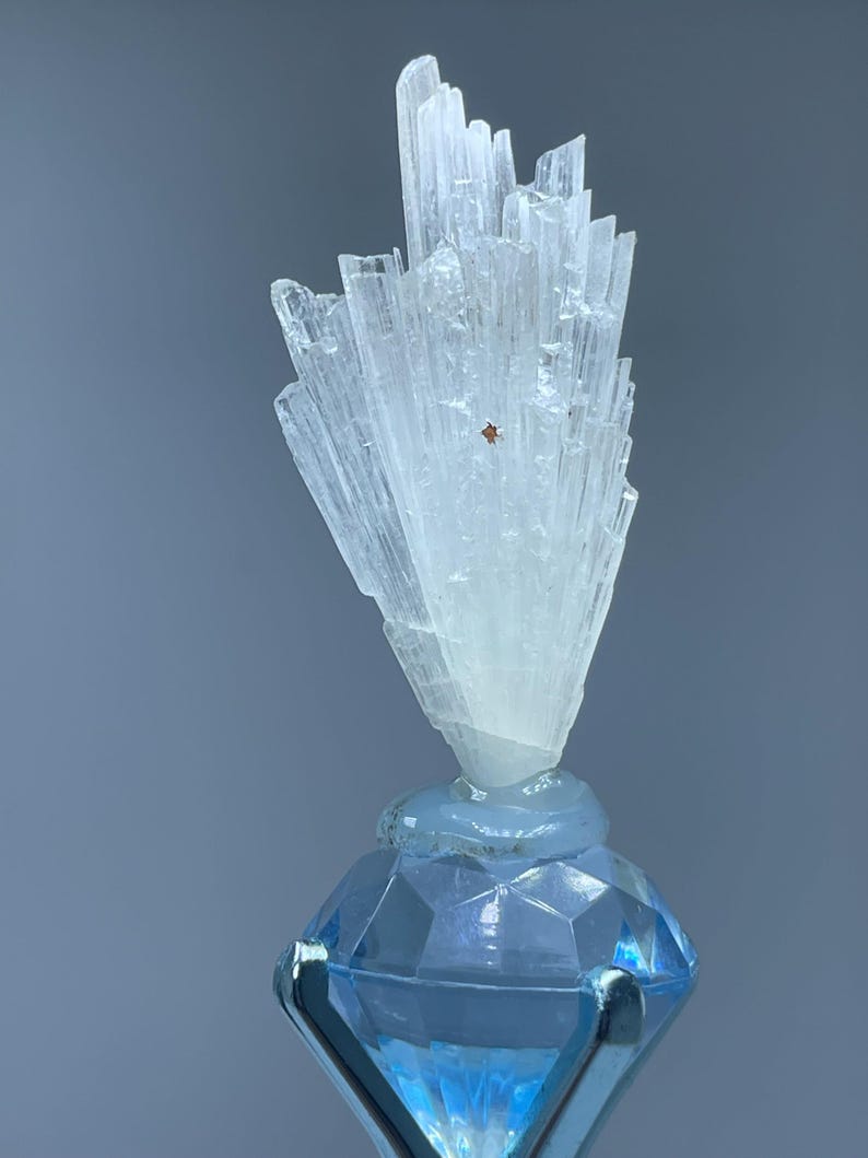 May include: A translucent, white, fan-shaped mineral cluster with vertical striations and a small brown inclusion, mounted atop a faceted, light blue, diamond-shaped glass base held by a silver-toned metal prong setting.