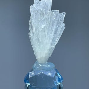 May include: A light blue crystal bottle stopper with a faceted diamond-shaped base and a cluster of clear, elongated crystals on top. The stopper is made of glass and has a silver-colored metal stand.
