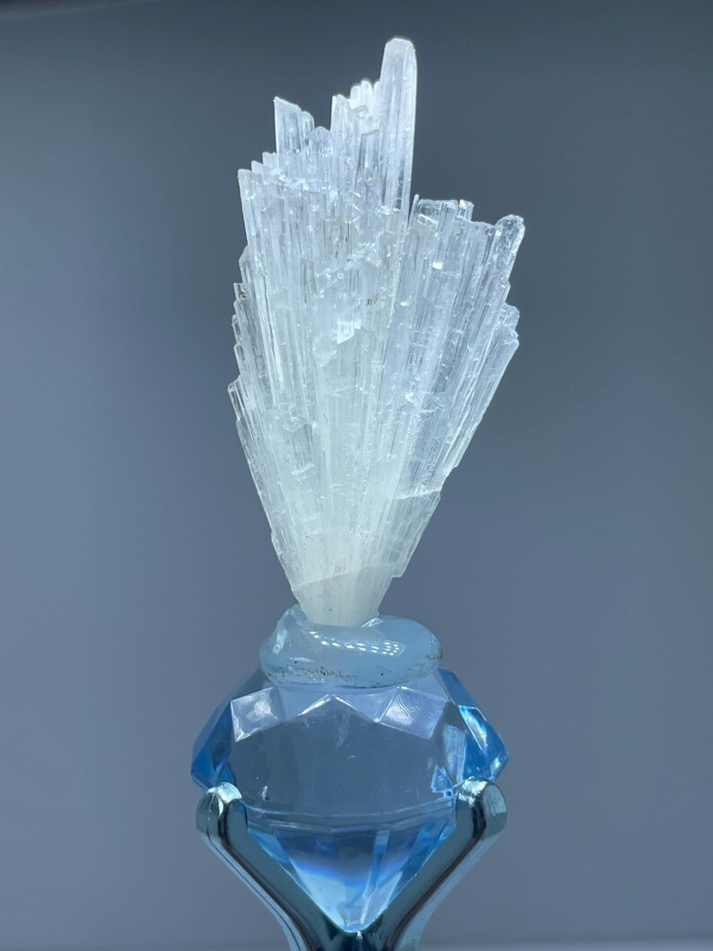 May include: A light blue crystal bottle stopper with a faceted diamond-shaped base and a cluster of clear, elongated crystals on top. The stopper is made of glass and has a silver-colored metal stand.