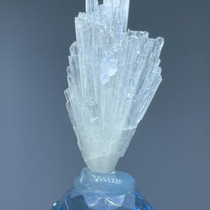 May include: A light blue crystal cluster with a fan-like shape, mounted on a faceted blue base. The crystal structure is composed of numerous, slender, translucent crystals. The base is a lighter shade of blue, with a geometric design.