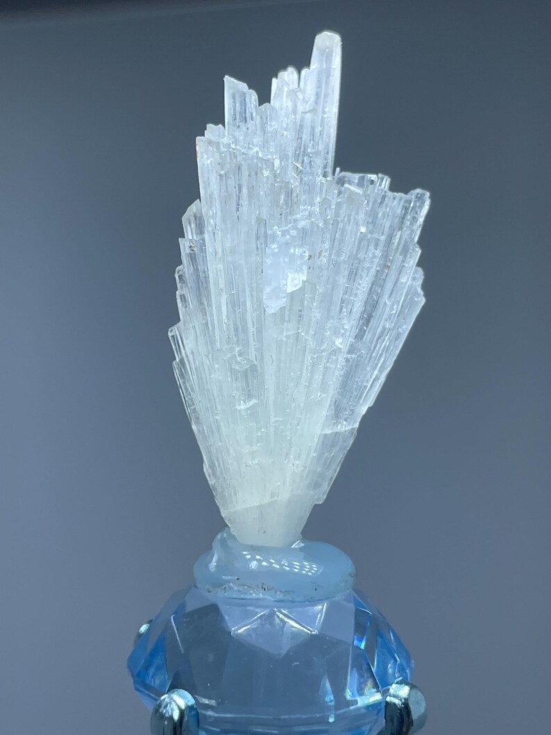 May include: A light blue crystal cluster with a fan-like shape, mounted on a faceted blue base. The crystal structure is composed of numerous, slender, translucent crystals. The base is a lighter shade of blue, with a geometric design.