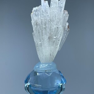 May include: A cluster of white, translucent crystals on a blue, diamond-shaped base. The crystals are arranged in a fan-like shape and have a natural, rough texture.