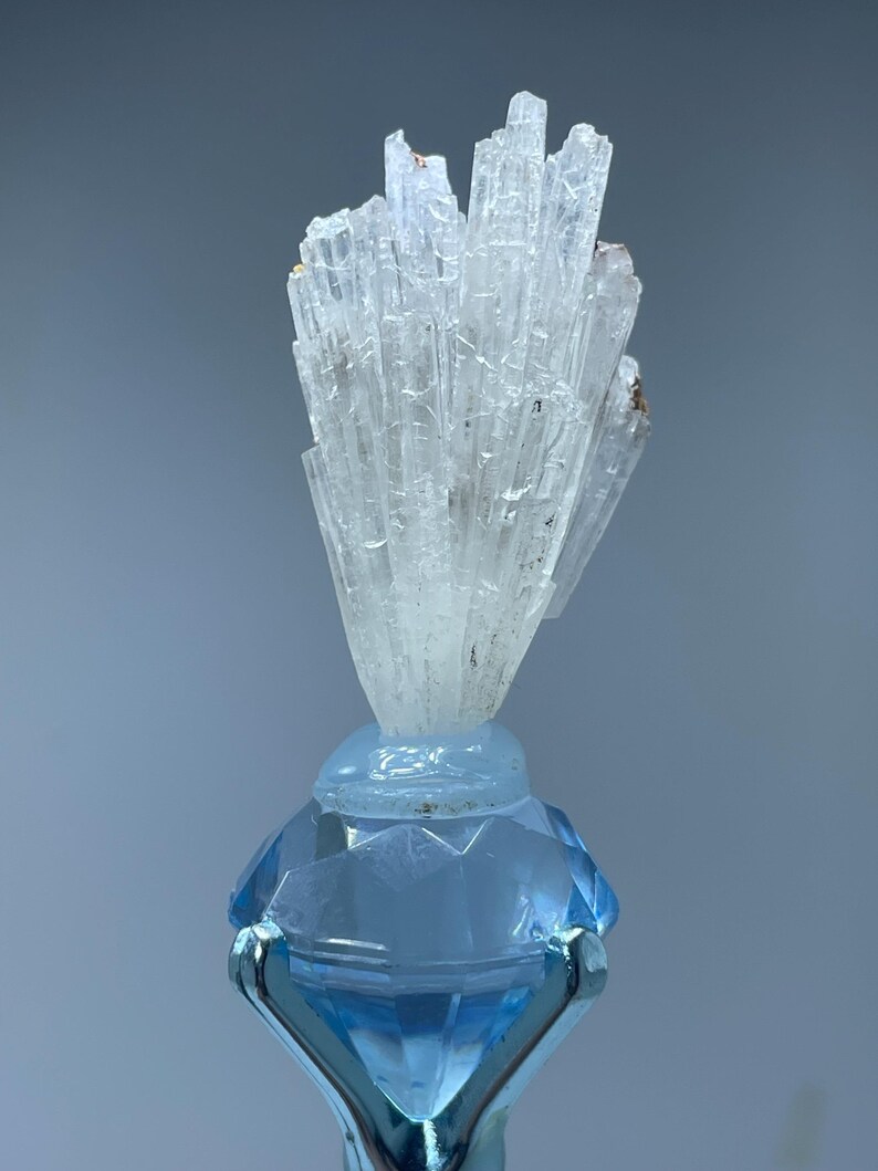 May include: A cluster of white, translucent crystals on a blue, diamond-shaped base. The crystals are arranged in a fan-like shape and have a natural, rough texture.