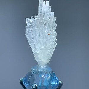 May include: A cluster of translucent, white, needle-like crystals stands vertically atop a faceted, light blue, diamond-shaped base. The crystal formation features a small, singular, reddish-brown speck embedded within its structure.