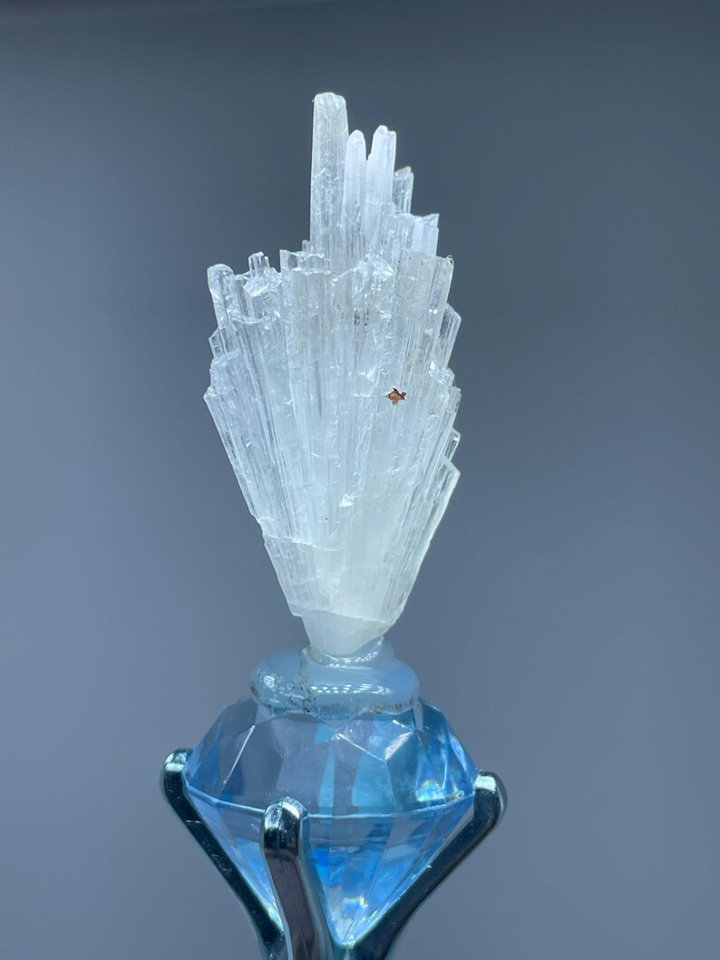 May include: A cluster of translucent, white, needle-like crystals stands vertically atop a faceted, light blue, diamond-shaped base. The crystal formation features a small, singular, reddish-brown speck embedded within its structure.