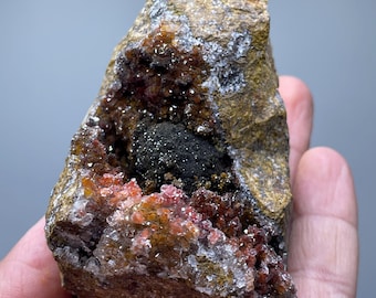 May include: A rough, natural rock formation with a cavity filled with sparkling crystals and clusters of reddish-orange and black minerals. The rock has a brown and tan exterior, and is held in a hand.