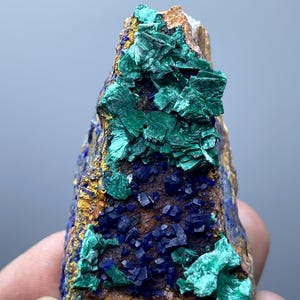 May include: A natural mineral specimen featuring a mix of colors. The rock has a brown base with patches of vibrant green and deep blue crystals. Yellow and gold accents are also visible, creating a striking contrast.