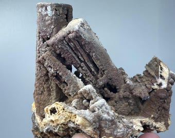 May include: A close-up of a natural mineral specimen. The specimen features a layered structure with a mix of brown, tan, and white colors. The texture appears rough and porous, with a crystalline appearance. The mineral is held in a hand.