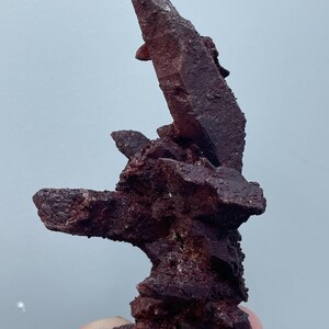 Könnte beinhalten: Ein Cluster aus tief roten Quarzkristallen mit einer rauen, strukturierten Oberfläche. Die Kristalle sind in einer vertikalen Formation angeordnet, wobei der höchste Kristall nach oben ragt.