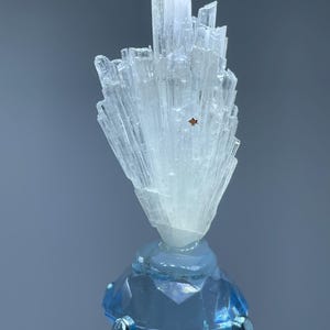 May include: A translucent white mineral specimen with elongated, needle-like crystal formations arranged in a fan shape, featuring a small reddish-brown square inclusion, mounted atop a faceted, light blue glass base with silver prongs.