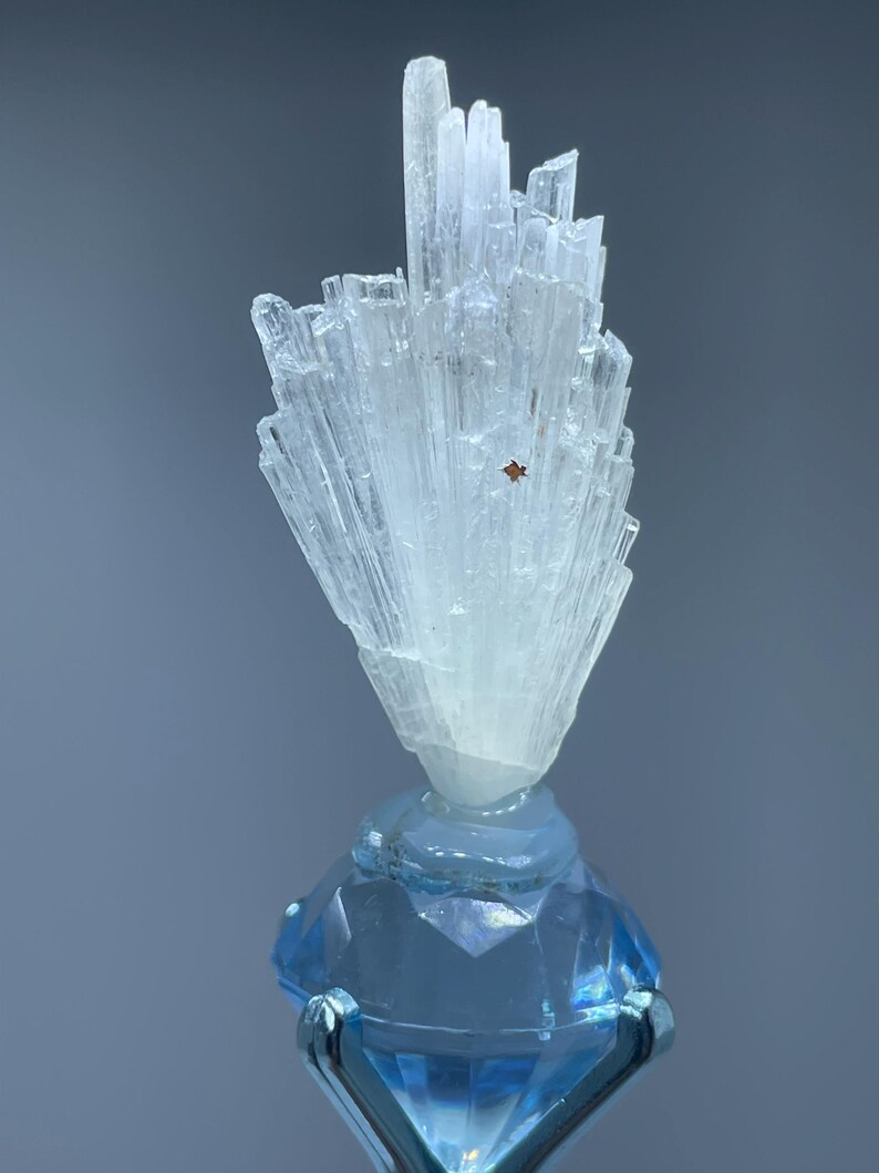 May include: A translucent white mineral specimen with elongated, needle-like crystal formations arranged in a fan shape, featuring a small reddish-brown square inclusion, mounted atop a faceted, light blue glass base with silver prongs.