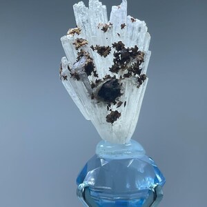 May include: A cluster of translucent, white, needle-like crystals featuring dark brown, metallic-looking inclusions, mounted atop a faceted, light blue, diamond-shaped glass base held by a silver-toned metal pronged stand.