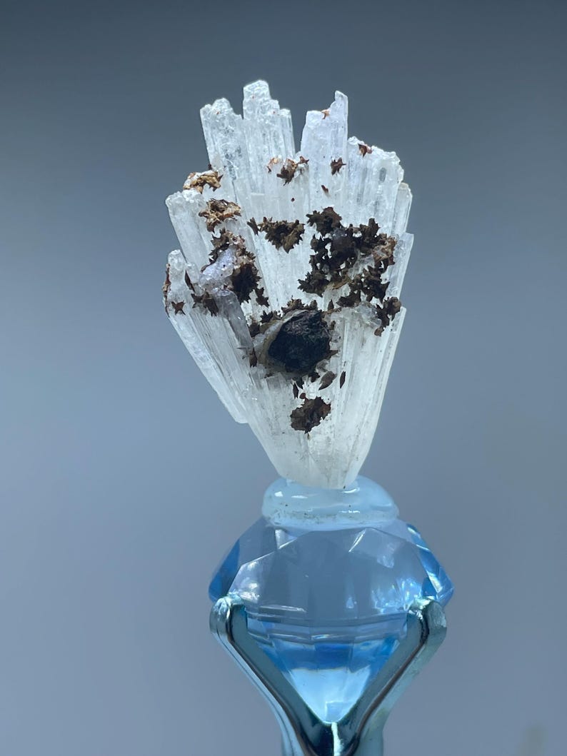 May include: A cluster of translucent, white, needle-like crystals featuring dark brown, metallic-looking inclusions, mounted atop a faceted, light blue, diamond-shaped glass base held by a silver-toned metal pronged stand.