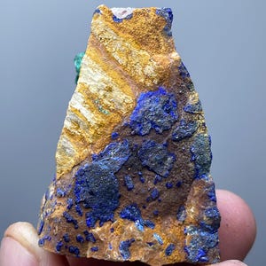 May include: A rough-cut mineral specimen featuring a mix of colors. The rock has a base of brown and tan hues, with striking patches of deep blue. The texture appears rough and natural, showcasing the mineral's raw beauty.