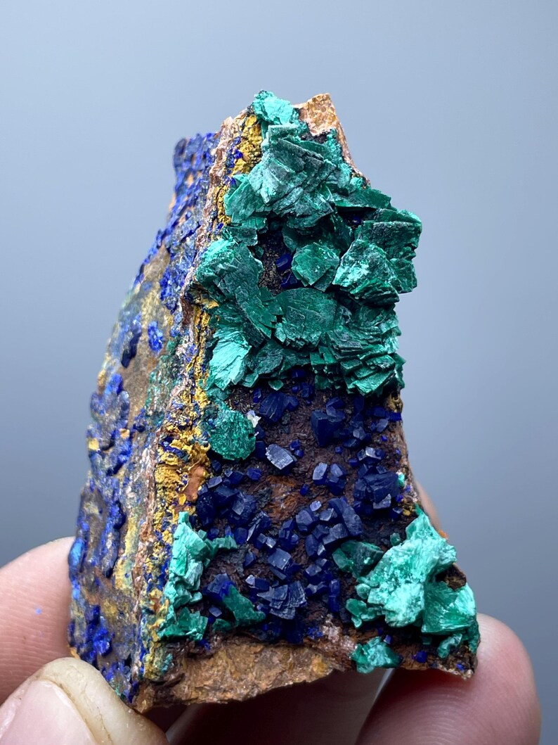 May include: A natural mineral specimen featuring a combination of vibrant colors. The piece showcases deep blue azurite crystals, contrasted by bright green malachite formations. The mineral is set against a backdrop of earthy brown and gold hues.