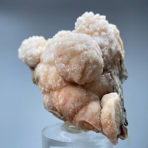 Könnte beinhalten: Eine Ansammlung von rosa und weißen Mineralformationen mit einer rauen, strukturierten Oberfläche. Das Mineralstück wird auf einem klaren Acrylsockel präsentiert.