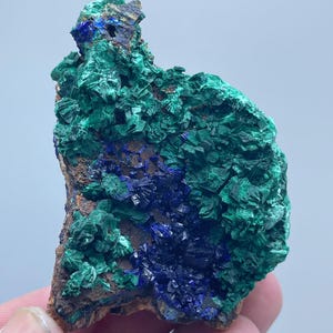 May include: A close-up of a mineral specimen featuring a combination of deep blue and vibrant green crystals. The mineral has a rough, textured surface with a mix of colors and shapes, set against a neutral background.
