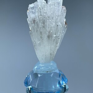 May include: A cluster of white selenite crystals on a blue diamond-shaped stand. The crystals are long and thin, and they are arranged in a fan shape.