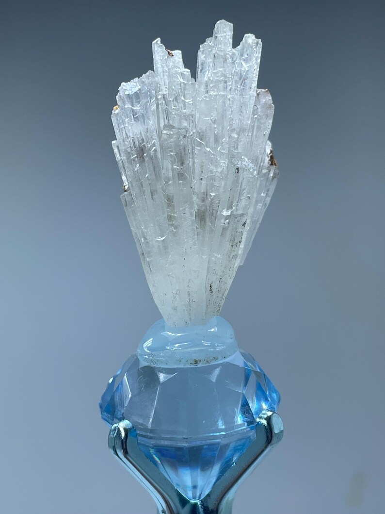 May include: A cluster of white selenite crystals on a blue diamond-shaped stand. The crystals are long and thin, and they are arranged in a fan shape.