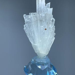 May include: A clear, crystalline mineral specimen with a cluster of elongated, light-colored crystals atop a faceted, light blue gemstone. The gemstone is held by a silver metal stand, creating a striking contrast.
