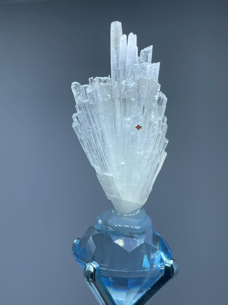 May include: A clear, crystalline mineral specimen with a cluster of elongated, light-colored crystals atop a faceted, light blue gemstone. The gemstone is held by a silver metal stand, creating a striking contrast.