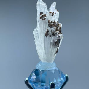 May include: A clear, crystalline mineral specimen with brown inclusions is mounted on a light blue, faceted gemstone. The gemstone is held in place by a silver metal setting. The background is a soft, muted blue.