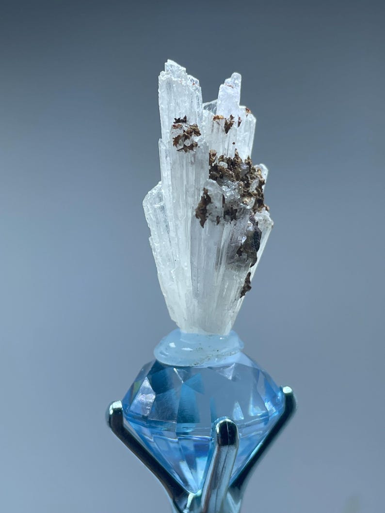 May include: A clear, crystalline mineral specimen with brown inclusions is mounted on a light blue, faceted gemstone. The gemstone is held in place by a silver metal setting. The background is a soft, muted blue.
