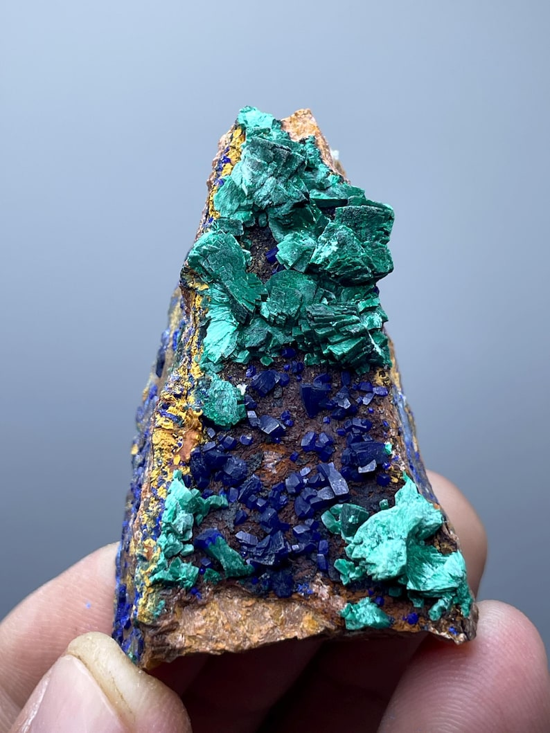 May include: A natural mineral specimen featuring a combination of colors and textures. The rock displays a mix of deep blue, vibrant green, and golden-brown hues. The surface is covered with crystalline formations.