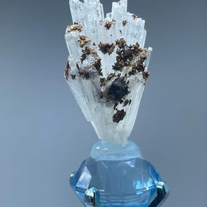 May include: A cluster of translucent, white, needle-like mineral crystals featuring dark brown, metallic-looking inclusions. The mineral specimen is mounted atop a faceted, light blue, diamond-shaped glass base held by a silver-toned metal prong.