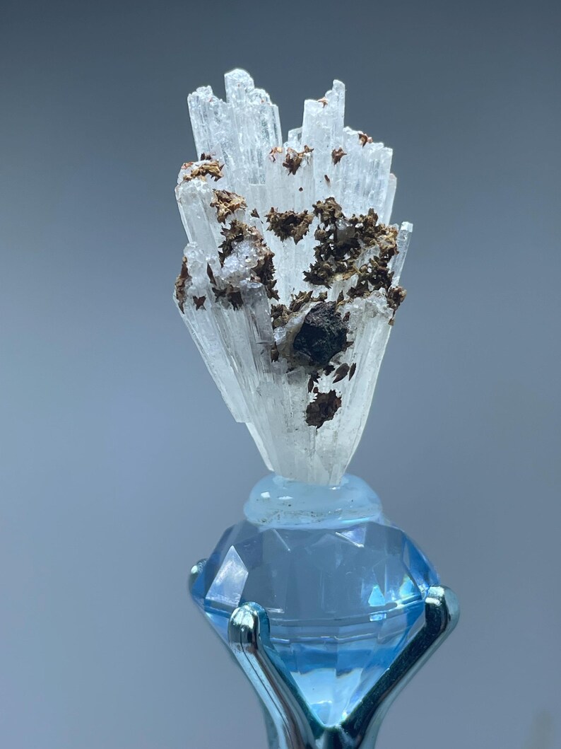 May include: A cluster of translucent, white, needle-like mineral crystals featuring dark brown, metallic-looking inclusions. The mineral specimen is mounted atop a faceted, light blue, diamond-shaped glass base held by a silver-toned metal prong.