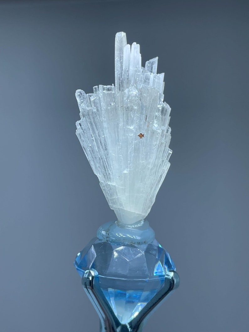 May include: A clear, crystalline mineral formation atop a light blue, faceted glass base. The mineral has a cluster of vertical, elongated crystals. The base is diamond-shaped with a silver metal support. The background is a solid, muted gray.