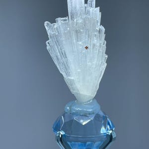 May include: A clear, crystalline mineral formation atop a faceted, light blue glass base. The mineral has a cluster of vertical, translucent crystals. The base is held by a silver-colored metal stand, creating a striking contrast.