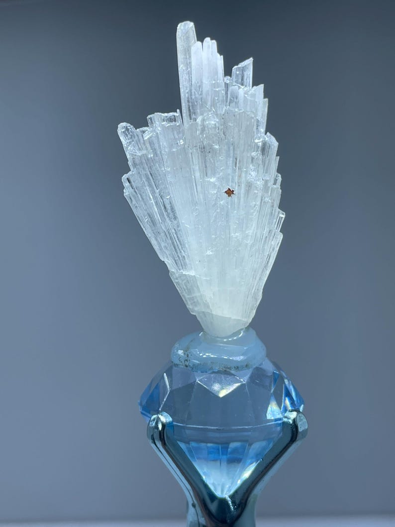 May include: A clear, crystalline mineral formation atop a faceted, light blue glass base. The mineral has a cluster of vertical, translucent crystals. The base is held by a silver-colored metal stand, creating a striking contrast.