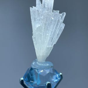 May include: A white selenite crystal cluster on a blue diamond-shaped stand. The crystal is a cluster of thin, needle-like crystals.