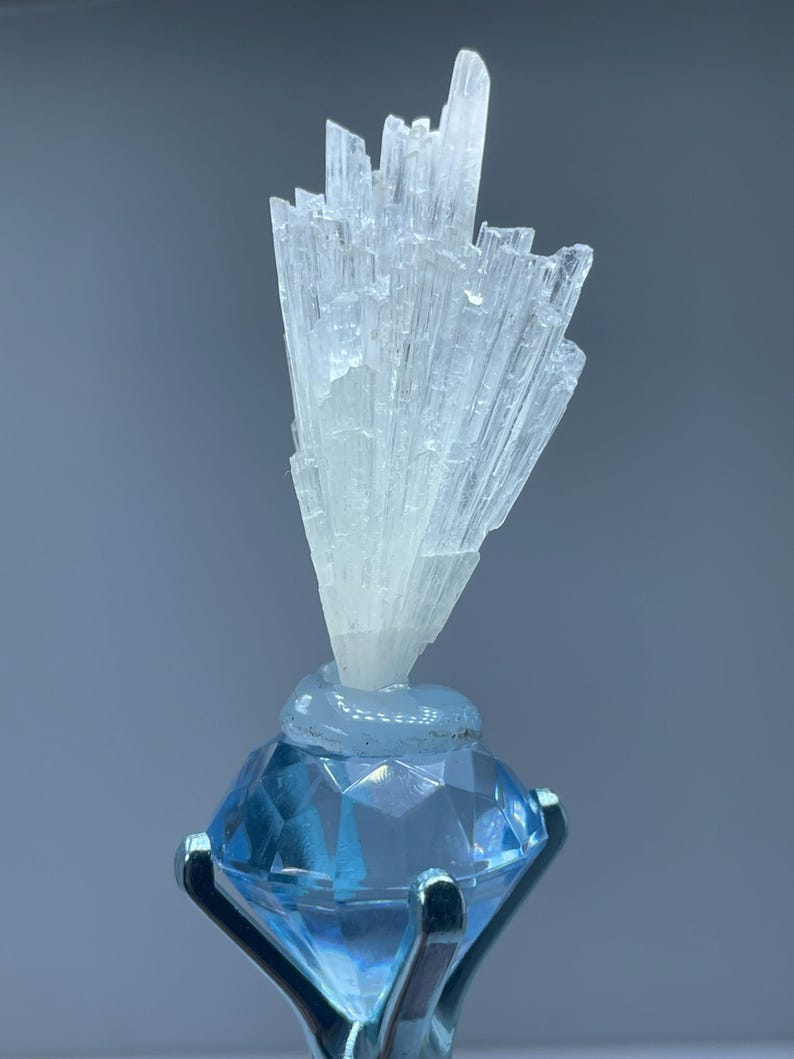 May include: A white selenite crystal cluster on a blue diamond-shaped stand. The crystal is a cluster of thin, needle-like crystals.