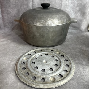 May include: A silver-toned aluminum Dutch oven with a matching lid featuring a black knob handle. The pot has two side handles and is accompanied by a circular, perforated metal steaming rack with a series of evenly spaced holes throughout.