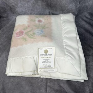 May include: A folded white baby blanket with a floral design in pink, blue, and green. The blanket has a satin border and a tag that reads "NORTH STAR BUTTERCUP" and the size is 42 x 60 inches.
