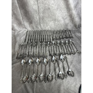 May include: A collection of vintage silverware, including knives, forks, and spoons, arranged on a gray surface. The silverware has ornate handles and a shiny, metallic finish. The set includes various sizes of forks, spoons, and knives.