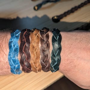 May include: A set of six braided leather bracelets in blue, brown, and tan colors. The bracelets are stacked on a person's wrist.