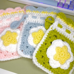 May include: Three rectangular crochet pouches featuring a scalloped top edge and a central white flower motif with a yellow star-shaped center. The pouches are colored in light pink, light blue, and olive green, each with a matching crochet drawstring.