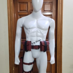 Könnte beinhalten: Ein dunkelrotes Gürtelsystem mit zwei rechteckigen Taschen, schwarzen Riemen und einer schwarzen Schnalle, das um die Taille getragen wird, ergänzt durch Beingurte und ein vertikales Holster. Der Text UK Props ist auf einem Etikett zu sehen.