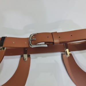 Puede incluir: Un cintur&oacute;n de cuero color canela con una hebilla de metal plateado y una trabilla de cuero a juego. El cintur&oacute;n incluye accesorios de metal plateado integrados que se conectan a dos paneles de cuero color canela curvados hacia abajo.