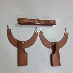 Puede incluir: Un conjunto de arn&eacute;s de cuero marr&oacute;n que consta de un cintur&oacute;n con hebilla plateada y dos piezas curvas para los hombros, cada una con un accesorio vertical estilo funda asegurado con remaches y clips de metal plateado.