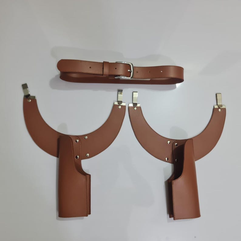 Puede incluir: Un conjunto de arn&eacute;s de cuero marr&oacute;n que consta de un cintur&oacute;n con hebilla plateada y dos piezas curvas para los hombros, cada una con un accesorio vertical estilo funda asegurado con remaches y clips de metal plateado.