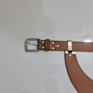 Puede incluir: Un cintur&oacute;n de cuero color canela con una hebilla de metal plateado y una trabilla de metal plateado a juego. El cintur&oacute;n presenta costuras visibles a lo largo de los bordes e incluye cuatro remaches de metal plateado cerca de la hebilla.