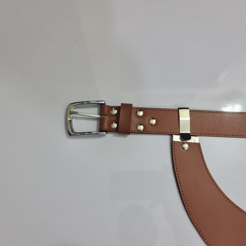 Puede incluir: Un cintur&oacute;n de cuero color canela con una hebilla de metal plateado y una trabilla de metal plateado a juego. El cintur&oacute;n presenta costuras visibles a lo largo de los bordes e incluye cuatro remaches de metal plateado cerca de la hebilla.