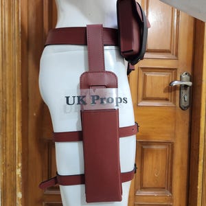 Puede incluir: Un conjunto de cinturón y funda para el muslo de color rojo oscuro, similar al cuero, con una bolsa rectangular vertical, dos correas ajustables para la pierna y una bolsa para la cadera. El texto "UK Props" aparece en una capa transparente.