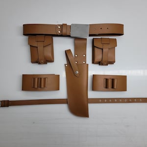 May include: A brown leather utility belt set with multiple pouches, a knife sheath, and a separate belt section. The belt has a silver buckle and is designed for carrying tools or accessories. The set is laid out on a white surface.