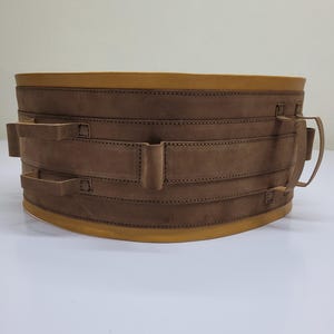 Könnte beinhalten: Ein brauner Ledergürtel mit mehreren Riemen und Schnallen. Der Gürtel hat einen gelben Rand an den Kanten.